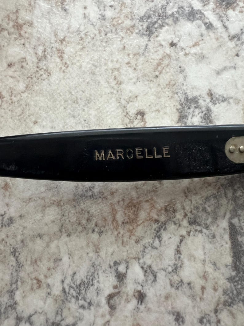 May include: Close-up of a black eyeglass frame with the name "MARCELLE" in silver lettering. The frame has a sleek, modern design. The background is a light-colored, speckled surface.