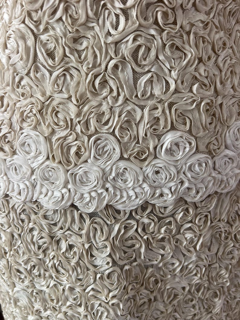 May include: A close-up of a white fabric with a textured pattern of small, raised rosettes. The fabric has a delicate, feminine look.
