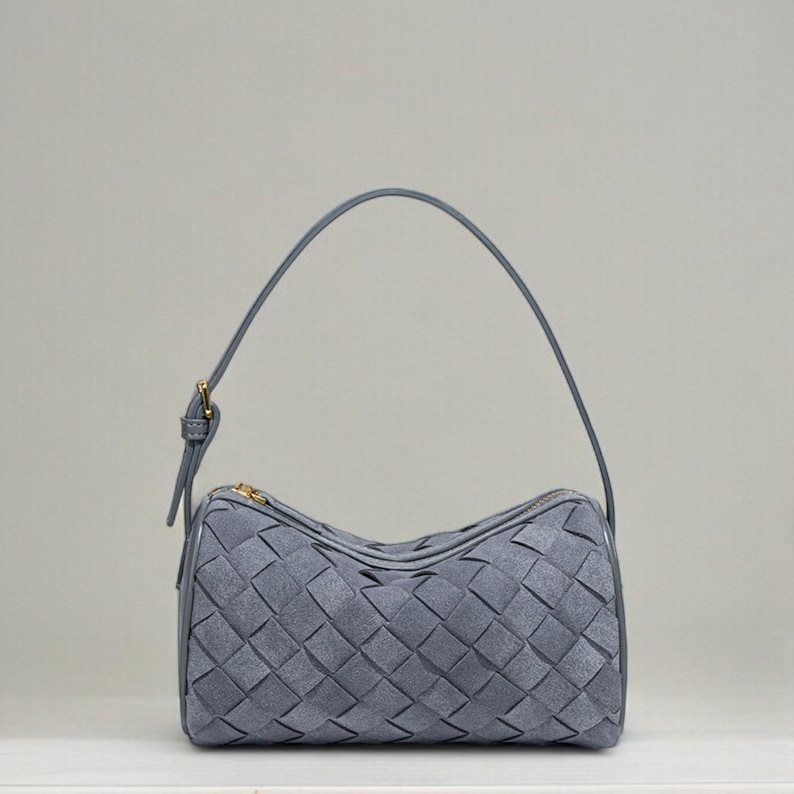 Puede incluir: Un bolso gris con dise&ntilde;o trenzado. El bolso tiene forma redondeada, cierre de cremallera y una fina correa para el hombro. Los herrajes son de color dorado.