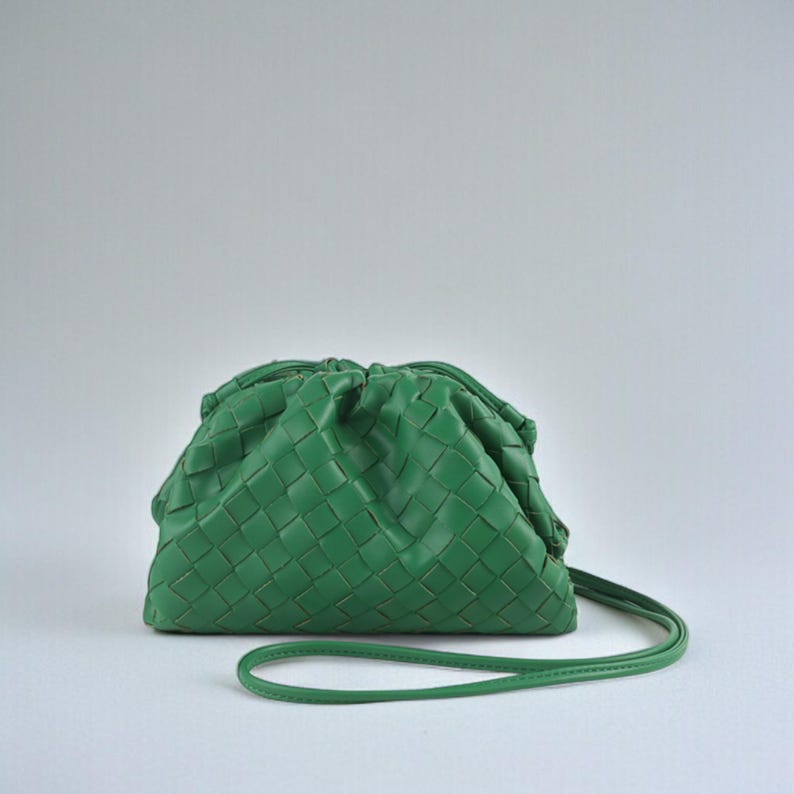 May include: A green woven leather clutch bag with a drawstring closure and a thin shoulder strap. The bag is a vibrant shade of green and has a soft, gathered appearance. The strap is approximately 100 cm long.