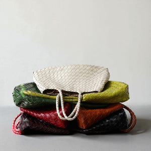 May include: A stack of woven leather tote bags in various colors, including white, green, red, brown, and black. The bags have braided handles and a textured, basket-weave design. The bags are stacked on top of each other.