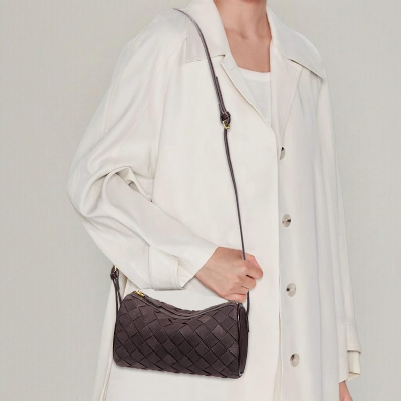 Puede incluir: Un bolso bandolera de cuero tejido marr&oacute;n oscuro con cierre de cremallera dorada. El bolso tiene forma cil&iacute;ndrica y una correa fina y ajustable. El bolso se lleva al hombro.