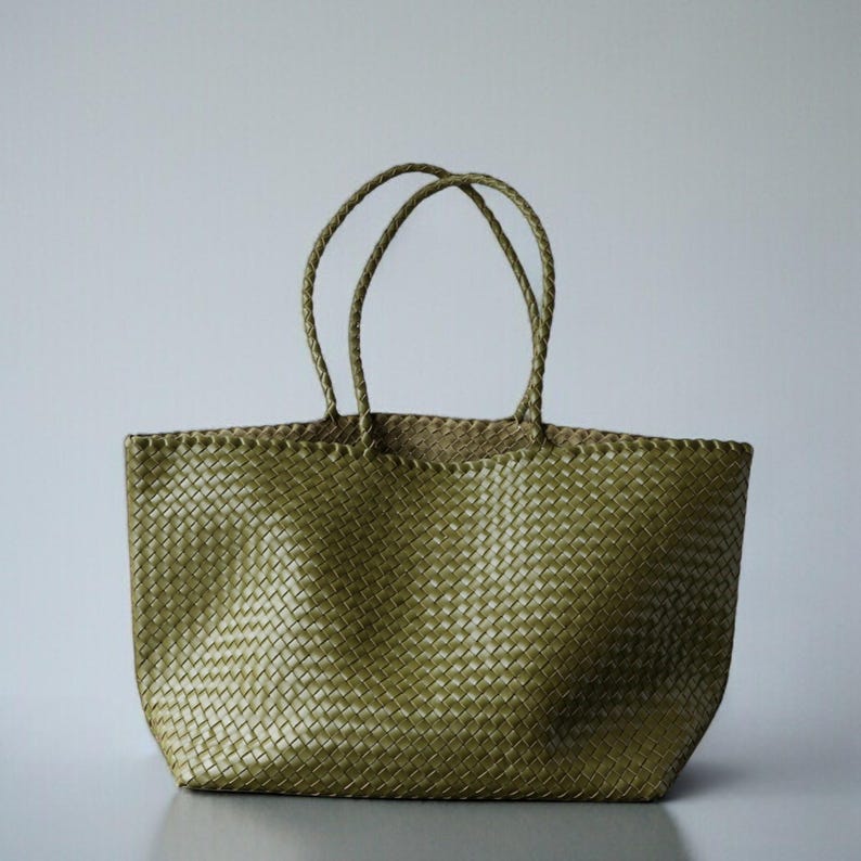 May include: A woven olive green tote bag with two braided handles. The bag has a rectangular shape and a textured surface. The handles are attached to the top of the bag and are made of the same material as the bag.