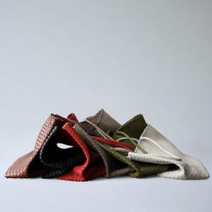 May include: A collection of woven leather tote bags in various colors, including brown, black, red, olive green, and gray. The bags are folded and stacked, showcasing their textured design and neutral color palette.