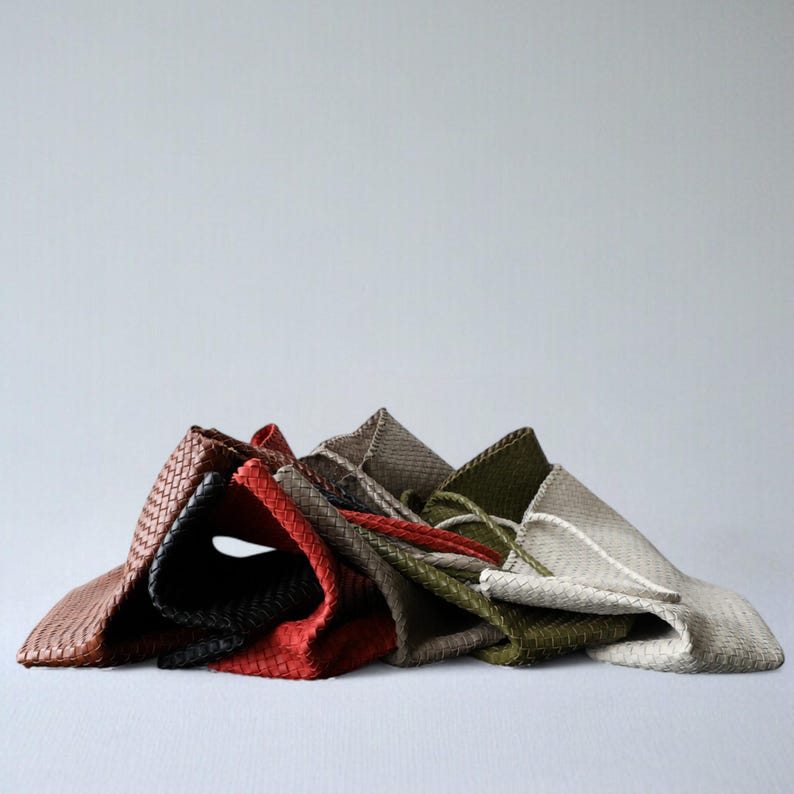 May include: A collection of woven leather tote bags in various colors, including brown, black, red, olive green, and gray. The bags are folded and stacked, showcasing their textured design and neutral color palette.
