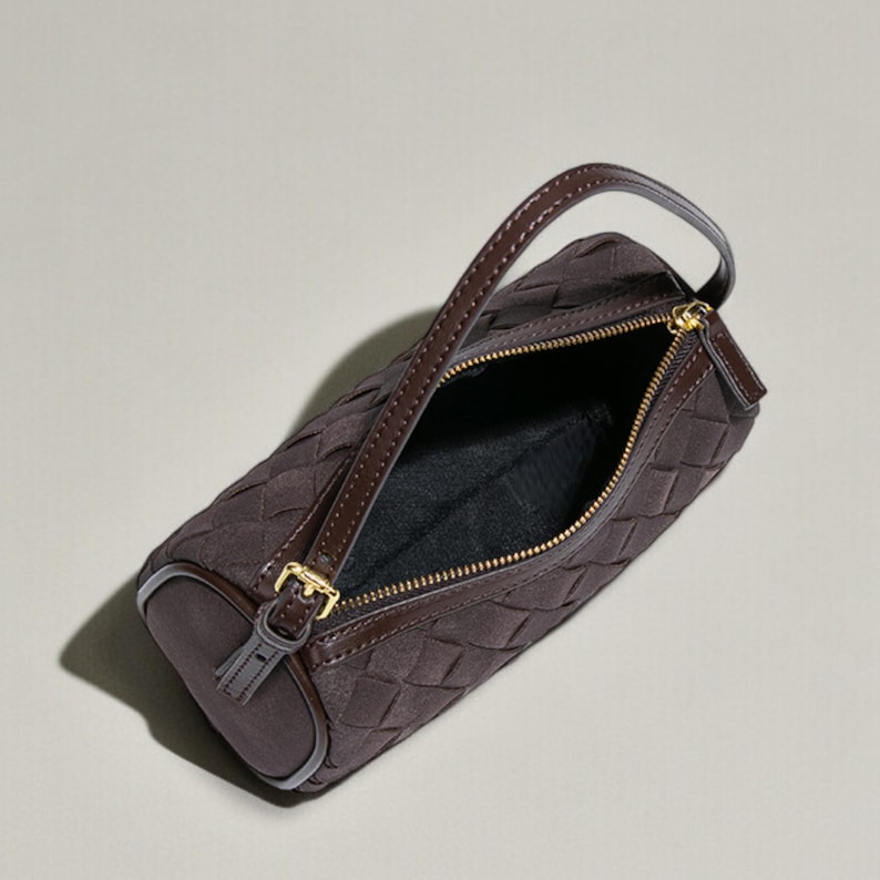 Puede incluir: Un bolso marr&oacute;n con un patr&oacute;n tejido y forma cil&iacute;ndrica. El bolso presenta un asa superior, una cremallera dorada y una hebilla dorada. El interior es negro. El bolso est&aacute; hecho de un material suave similar al ante.