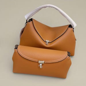 Pu&ograve; includere: Due borse in pelle color cuoio. La borsa pi&ugrave; grande ha una parte superiore curva con un manico bianco e una chiusura color argento. La borsa pi&ugrave; piccola &egrave; rettangolare con una patta curva e una chiusura color argento. Entrambe le borse hanno cuciture bianche.
