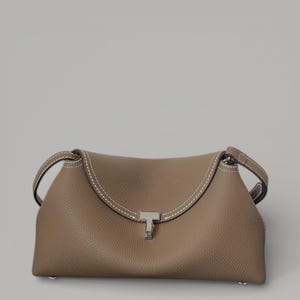 Pu&ograve; includere: Una borsa color talpa con una parte superiore curva e due piccole maniglie. La borsa presenta cuciture bianche lungo la parte superiore e una chiusura color argento. La borsa &egrave; realizzata in un materiale strutturato.