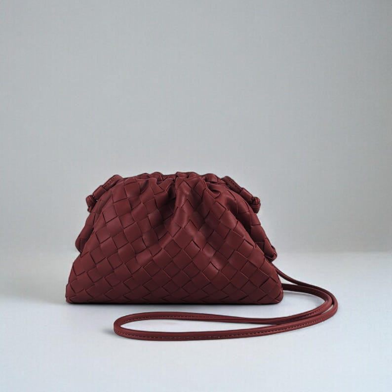 May include: A burgundy clutch bag with a woven pattern. The bag features a gathered top and a thin, matching strap. The material appears soft. Ideal for carrying small essentials.