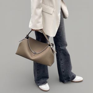 Pu&ograve; includere: Una borsa in pelle color talpa con patta superiore curva e chiusura color argento. La borsa ha un manico superiore e una tracolla. Le cuciture bianche accentuano i bordi. La borsa &egrave; tenuta da una persona che indossa un blazer bianco e jeans scuri.