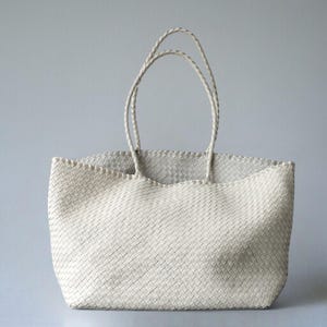 May include: Off-white woven tote bag with braided handles. The bag has a rectangular shape with a wide opening and a textured, basket-weave design. The handles are long enough to be worn over the shoulder.