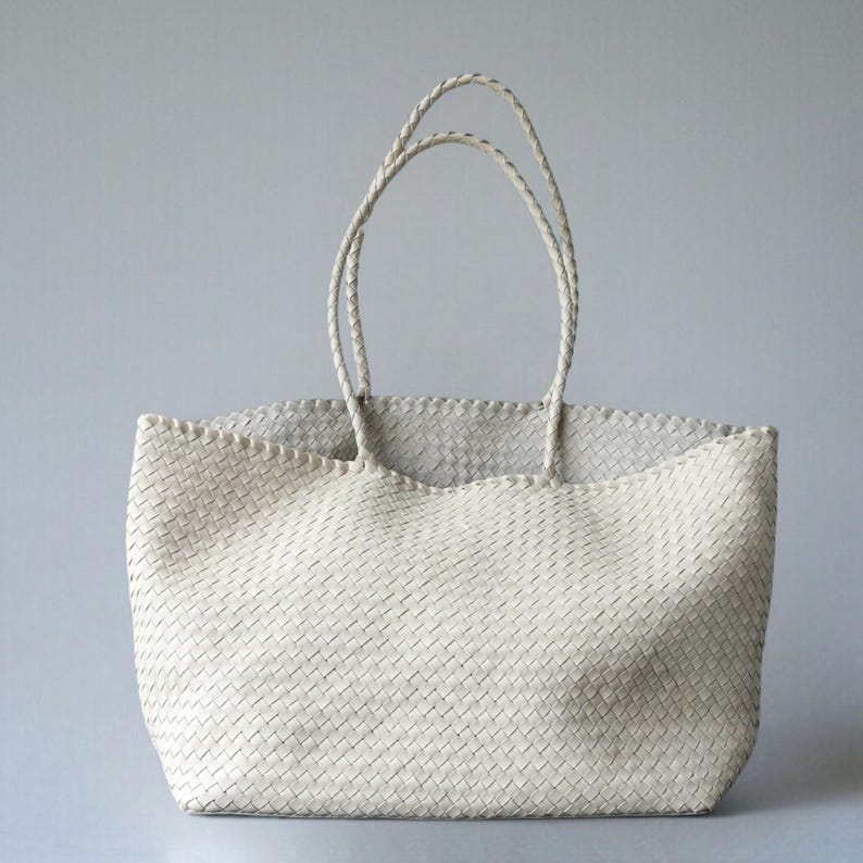 May include: Off-white woven tote bag with braided handles. The bag has a rectangular shape with a wide opening and a textured, basket-weave design. The handles are long enough to be worn over the shoulder.