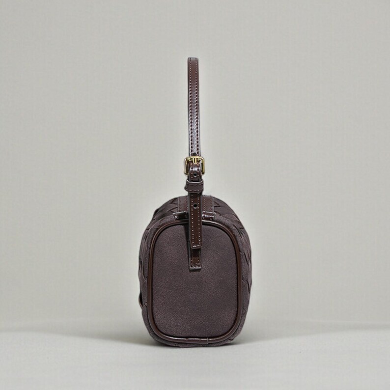 Puede incluir: Un bolso marr&oacute;n oscuro con un patr&oacute;n tejido y un asa corta. El bolso tiene forma rectangular con bordes redondeados y una hebilla dorada. El asa est&aacute; sujeta a la parte superior del bolso con una correa de cuero.