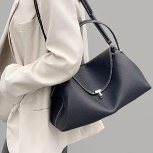 Pu&ograve; includere: Una borsa in pelle nera con cuciture bianche e una chiusura color argento. La borsa ha una forma strutturata con un manico superiore e una tracolla. La borsa &egrave; indossata da una persona che indossa un blazer color crema.