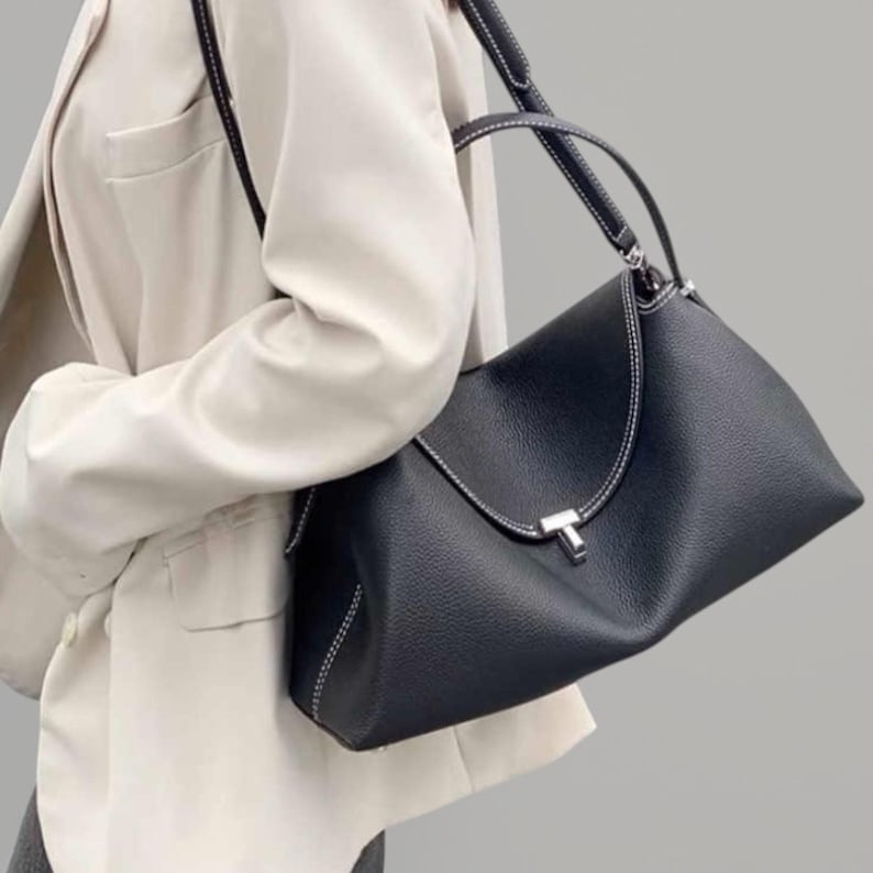 Pu&ograve; includere: Una borsa in pelle nera con cuciture bianche e una chiusura color argento. La borsa ha una forma strutturata con un manico superiore e una tracolla. La borsa &egrave; indossata da una persona che indossa un blazer color crema.