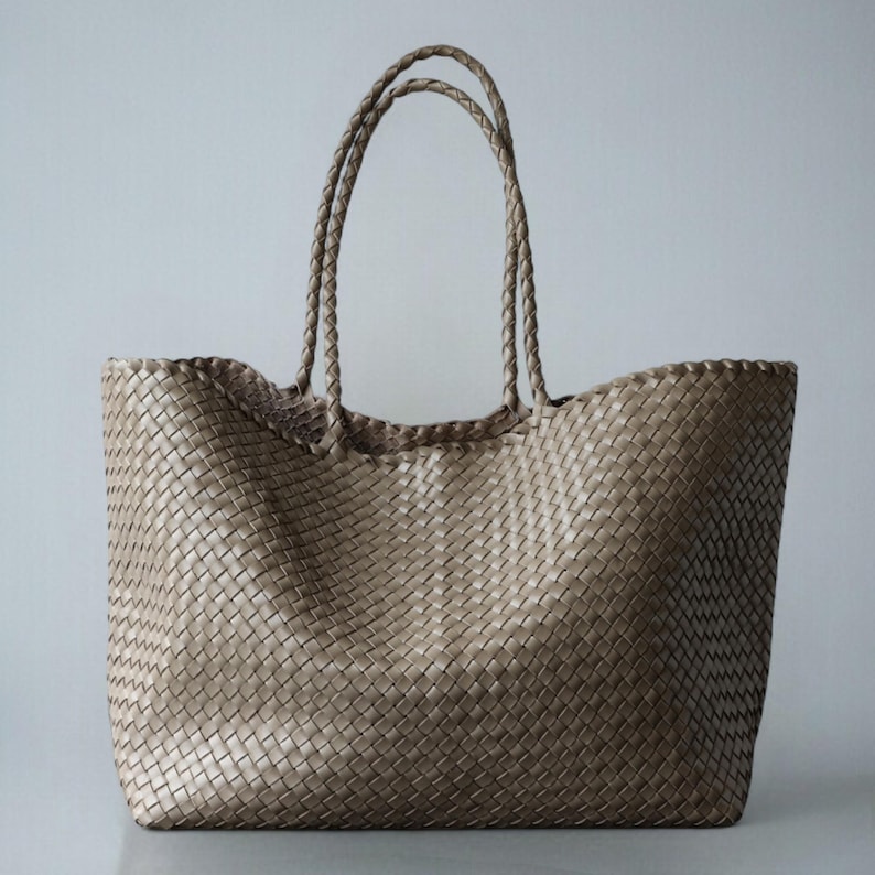 May include: A large, woven tote bag in a neutral beige color. The bag features two braided handles and a spacious interior. The woven pattern creates a textured surface, and the bag is designed for carrying items.