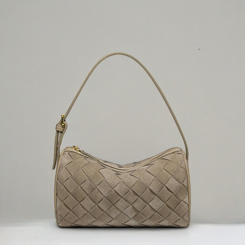 Puede incluir: Un bolso beige con un dise&ntilde;o tejido. El bolso tiene forma cil&iacute;ndrica con un asa superior y una cremallera dorada. El patr&oacute;n tejido est&aacute; hecho de piezas rectangulares de material.