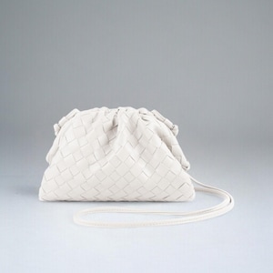 May include: Cream-coloured woven clutch bag with a long, thin strap. The bag features a soft, gathered design and a rectangular woven pattern. The strap matches the bag's colour. The bag is set against a white backdrop.