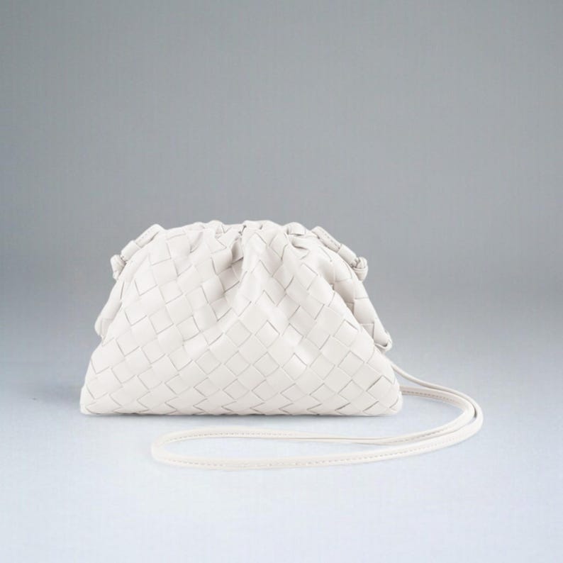 May include: Cream-coloured woven clutch bag with a long, thin strap. The bag features a soft, gathered design and a rectangular woven pattern. The strap matches the bag's colour. The bag is set against a white backdrop.