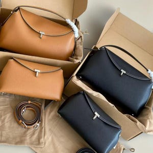 Pu&ograve; includere: Diverse borse in pelle marrone e nera sono esposte in scatole di cartone aperte. Le borse hanno una chiusura a patta con chiusura color argento e cuciture bianche. Sono visibili anche una tracolla e un sacchetto.