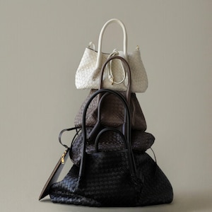 Può includere: Una pila di borse in pelle intrecciata in vari colori. Le borse includono una borsa tote bianca, marrone e nera, ciascuna con un design del manico distintivo. Le borse sono disposte verticalmente, mostrando le loro trame e forme.