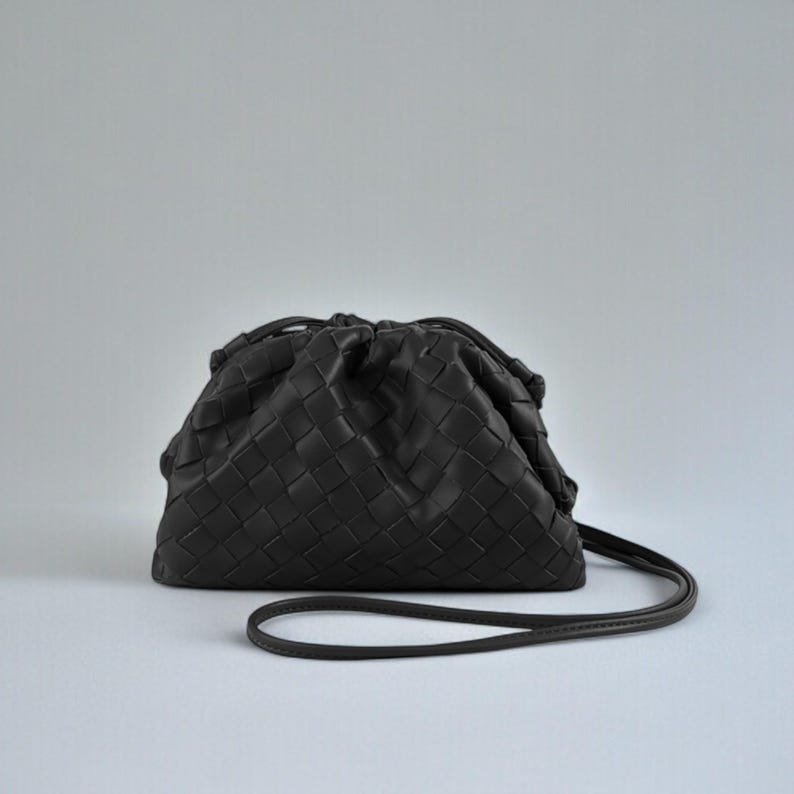 May include: A dark grey woven leather clutch bag with a drawstring closure and a thin shoulder strap. The bag has a rounded shape and a textured surface. The strap is approximately 100 cm long.