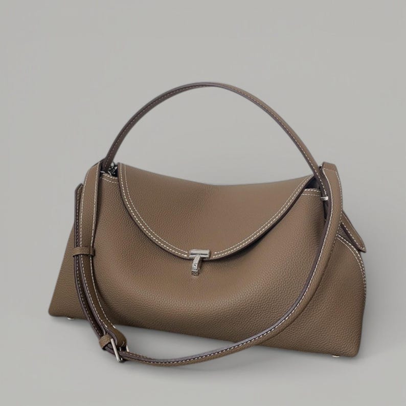 Pu&ograve; includere: Borsa a mano color talpa con patta superiore curva e chiusura argentata. Presenta un manico superiore e una tracolla staccabile. Le cuciture bianche accentuano i bordi, creando un contrasto. Realizzata in materiale strutturato.