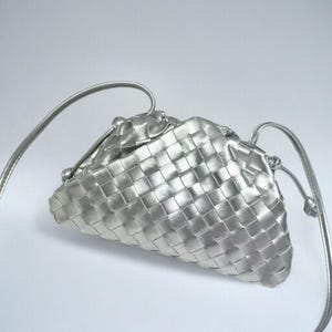May include: A silver-coloured, woven leather clutch bag with a shoulder strap. The bag has a triangular shape and a drawstring closure. The woven pattern creates a textured surface. The strap is thin and appears to be made of the same material as the bag.