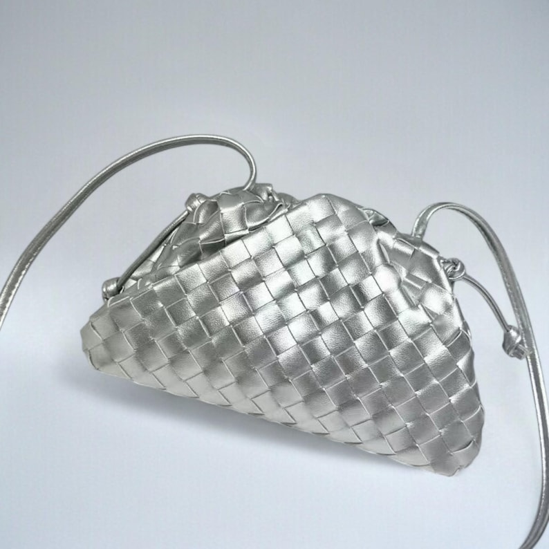 May include: A silver-coloured, woven leather clutch bag with a shoulder strap. The bag has a triangular shape and a drawstring closure. The woven pattern creates a textured surface. The strap is thin and appears to be made of the same material as the bag.