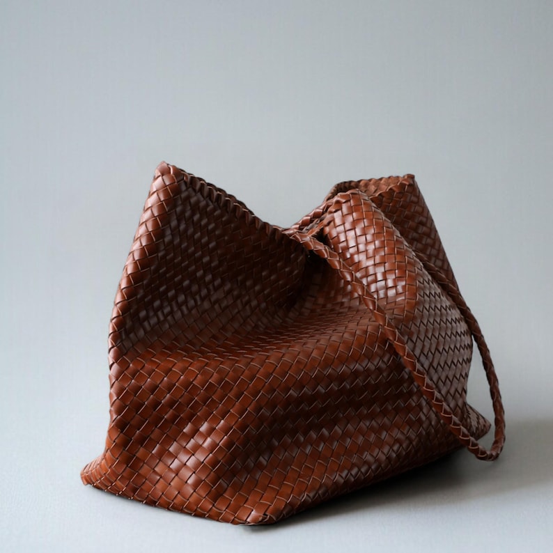 May include: A brown woven leather tote bag with a braided handle. The bag has a large, open design and a textured surface. The bag is made of a rich, warm brown leather.