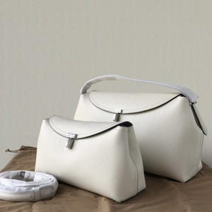 Pu&ograve; includere: Due borse in pelle bianca di diverse dimensioni. Le borse hanno una chiusura a patta curva con una chiusura color argento. Una borsa ha un manico superiore e l'altra una tracolla staccabile. Le borse sono su una superficie marrone.