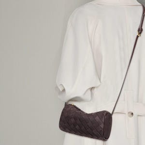 Puede incluir: Bolso bandolera de cuero tejido marr&oacute;n oscuro con forma cil&iacute;ndrica. El bolso tiene una correa larga y fina y una cremallera dorada. El bolso se lleva sobre un abrigo blanco.