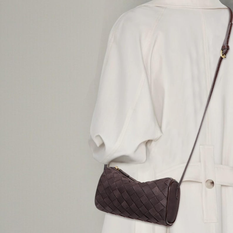 Puede incluir: Bolso bandolera de cuero tejido marr&oacute;n oscuro con forma cil&iacute;ndrica. El bolso tiene una correa larga y fina y una cremallera dorada. El bolso se lleva sobre un abrigo blanco.