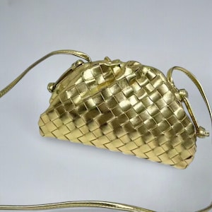 May include: A gold-coloured woven clutch bag with a long, thin strap. The bag has a rounded shape and a clasp closure. The woven pattern creates a textured surface. The strap is also gold and appears to be made of a similar material.