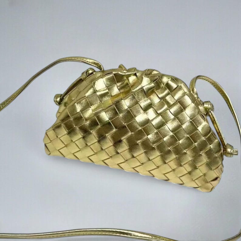May include: A gold-coloured woven clutch bag with a long, thin strap. The bag has a rounded shape and a clasp closure. The woven pattern creates a textured surface. The strap is also gold and appears to be made of a similar material.