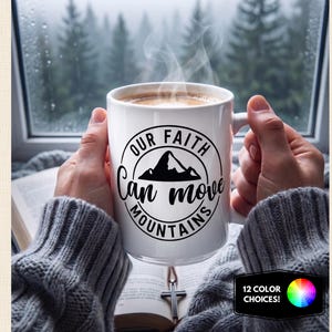 May include: A white ceramic mug featuring a black circular graphic with a mountain illustration and the text Our Faith Can Move Mountains. The mug is held by hands wearing a gray cable knit sweater, with steam rising from the hot beverage inside.