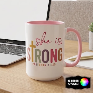 May include: A white ceramic mug with a pink interior and handle. The front features the text "she is" in pink script, "STRONG" in large, multi-colored block letters with a cross replacing the "T", and "PROVERBS 31:25" in small text.