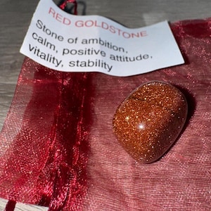 May include: A polished, reddish-brown Red Goldstone gemstone. The stone is smooth and oval-shaped, with a sparkling, glittery appearance. A small tag reads "Stone of ambition, calm, positive attitude, vitality, stability."
