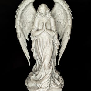 May include: A detailed statue of an angel with outstretched wings, hands clasped in prayer, and flowing robes. The sculpture is a light gray color and stands on a textured base. The background is solid black.