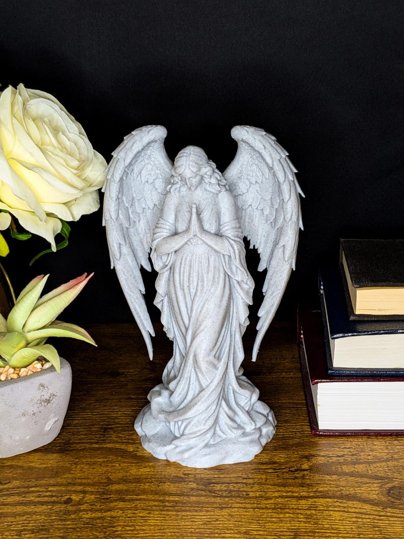 May include: A detailed, gray angel statue with outstretched wings and clasped hands. The angel is wearing a flowing gown and stands on a textured base. The statue is set against a dark background, with a white rose and books nearby.