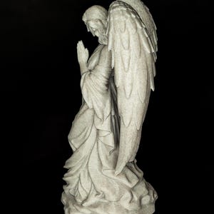 May include: A detailed statue of an angel in a praying pose. The angel is depicted with large, detailed wings and flowing robes. The statue is a light gray color and stands on a textured base. The background is black.