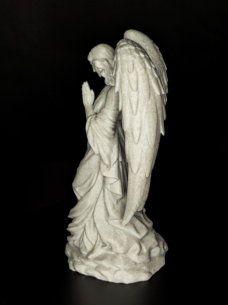 May include: A detailed statue of an angel in a praying pose. The angel is depicted with large, detailed wings and flowing robes. The statue is a light gray color and stands on a textured base. The background is black.