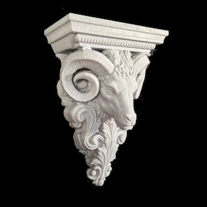 May include: A light grey wall corbel featuring a sculpted ram head with prominent curled horns. The architectural support piece includes intricate acanthus leaf carvings extending downwards from the ram, finished with a flat rectangular top shelf.