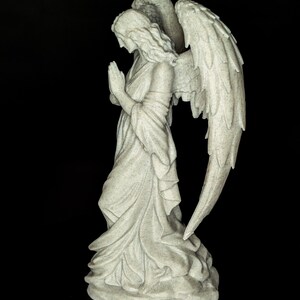 May include: A stone-colored angel statue with folded hands in prayer. The angel wears a flowing gown and has large, detailed wings. The statue is set against a black background.