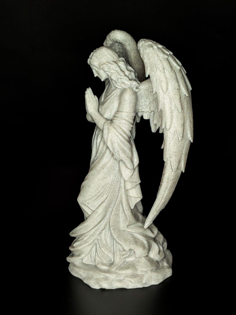 May include: A stone-colored angel statue with folded hands in prayer. The angel wears a flowing gown and has large, detailed wings. The statue is set against a black background.