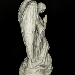 May include: A stone-colored angel statue with large, detailed wings and a serene expression. The angel is kneeling with hands clasped in prayer, wearing a flowing robe. The statue is set against a black background.