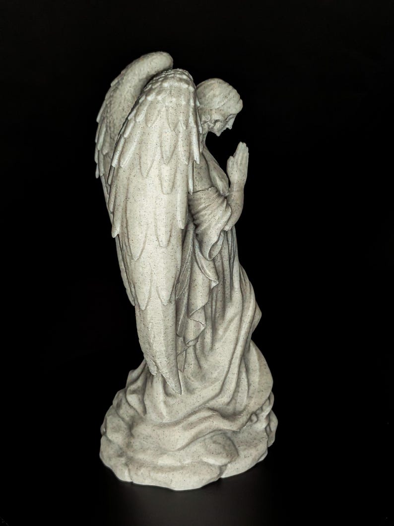 May include: A stone-colored angel statue with large, detailed wings and a serene expression. The angel is kneeling with hands clasped in prayer, wearing a flowing robe. The statue is set against a black background.