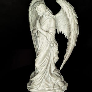 May include: A detailed statue of an angel with large, textured wings. The angel is depicted in a flowing gown, with hands clasped in prayer. The statue is a light gray color and stands on a textured base, set against a black background.