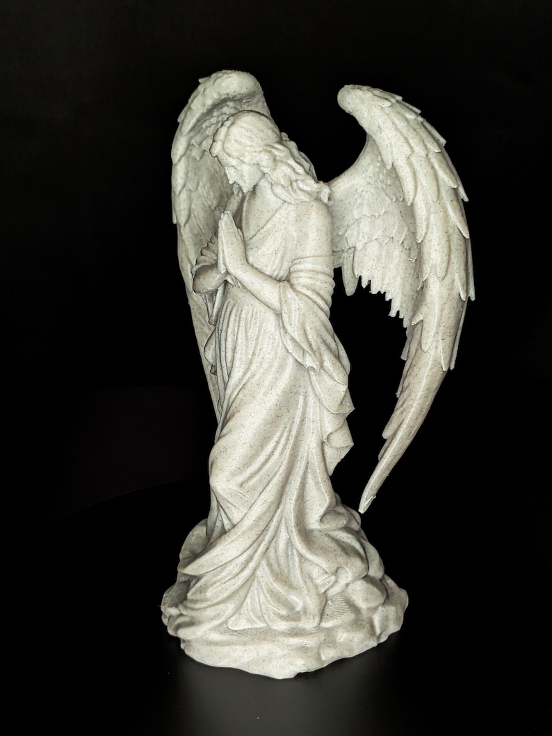 May include: A detailed statue of an angel with large, textured wings. The angel is depicted in a flowing gown, with hands clasped in prayer. The statue is a light gray color and stands on a textured base, set against a black background.