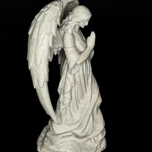 May include: A stone-colored angel statue with large, detailed wings. The angel is depicted in a praying pose, wearing a flowing gown. The statue has a textured surface and is set against a black background.
